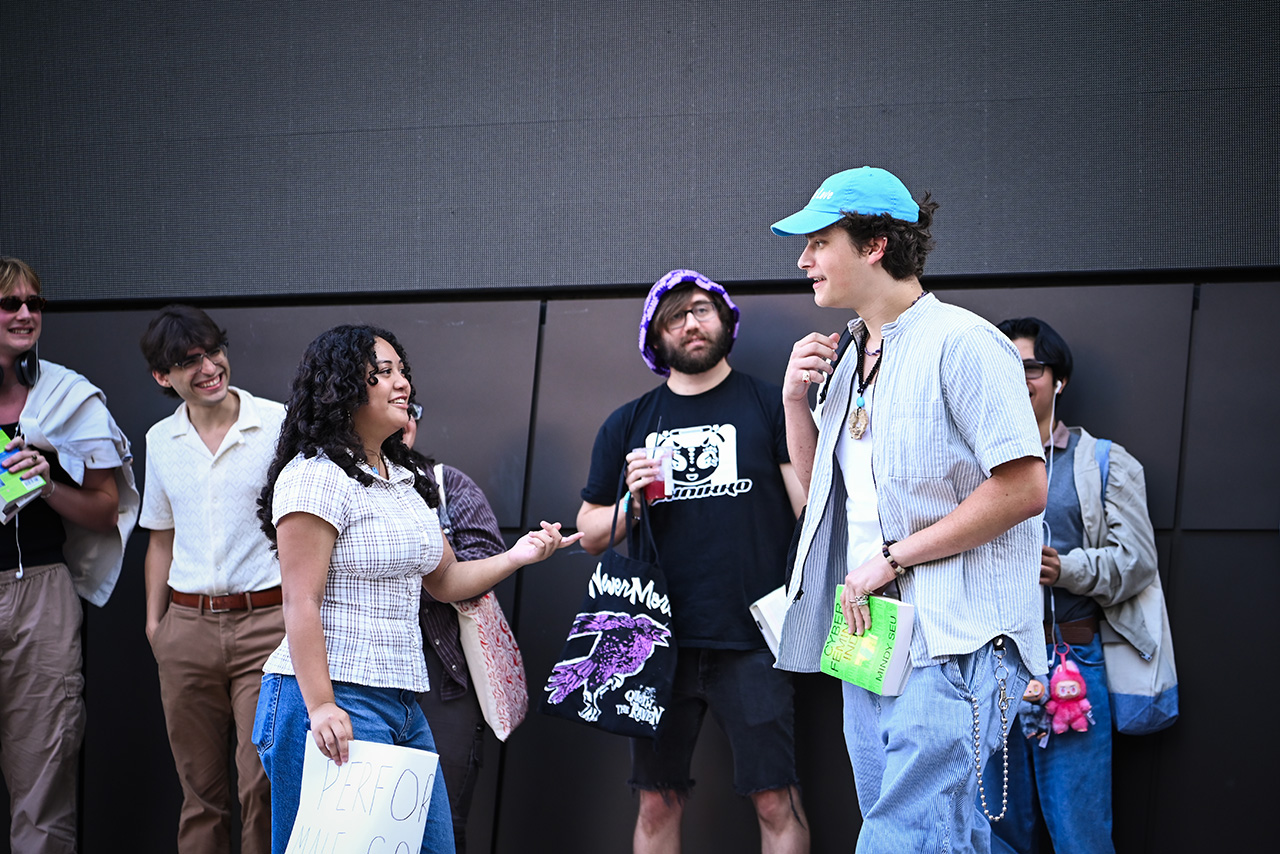 Photo of a young woman with blouse talking to a male contestor with blue cap and white shirt during the Boston Performative Male Contest