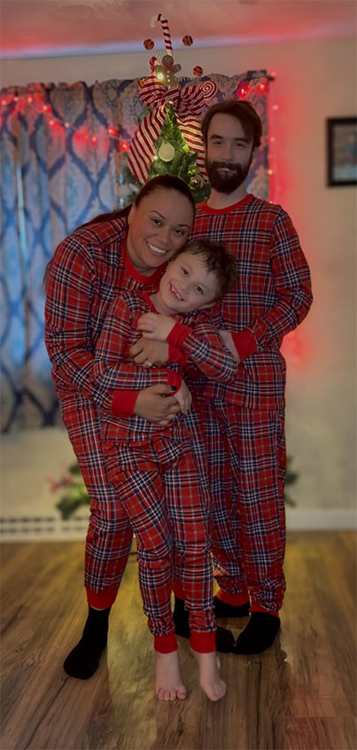 A smiling family of three wearing matching red plaid pajamas poses together in a cozy living room. The young boy is being hugged from behind by the woman while the man stands close behind them, all in front of a small decorated Christmas tree with holiday lights in the background.