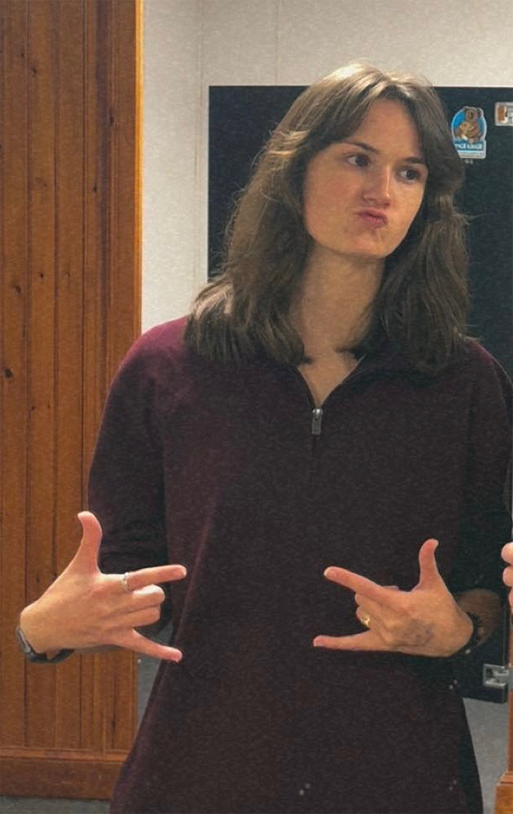 A young woman with shoulder-length brown hair poses playfully, displaying both hands in a casual gesture. She is wearing a maroon zip-up top and stands in front of a wooden wall in an indoor setting.
