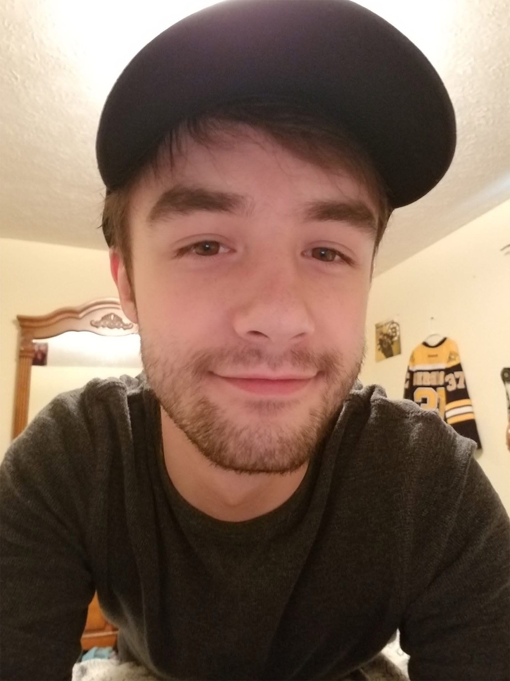 A young person with short brown hair and a thin beard wearing a black baseball hat smiles slightly at the camera in a warmly lit bedroom. A sports jersey hangs on the wall in the background.
