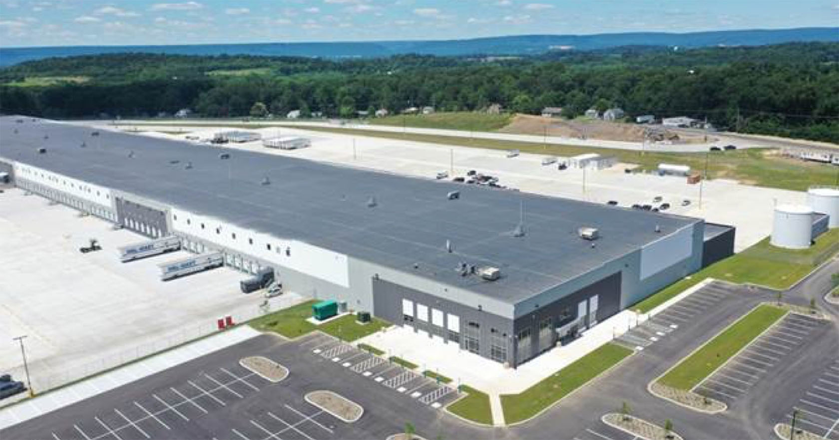 A large industrial warehouse complex with multiple loading docks and semi-trailers parked outside, surrounded by a wide parking area and green fields under a blue sky.