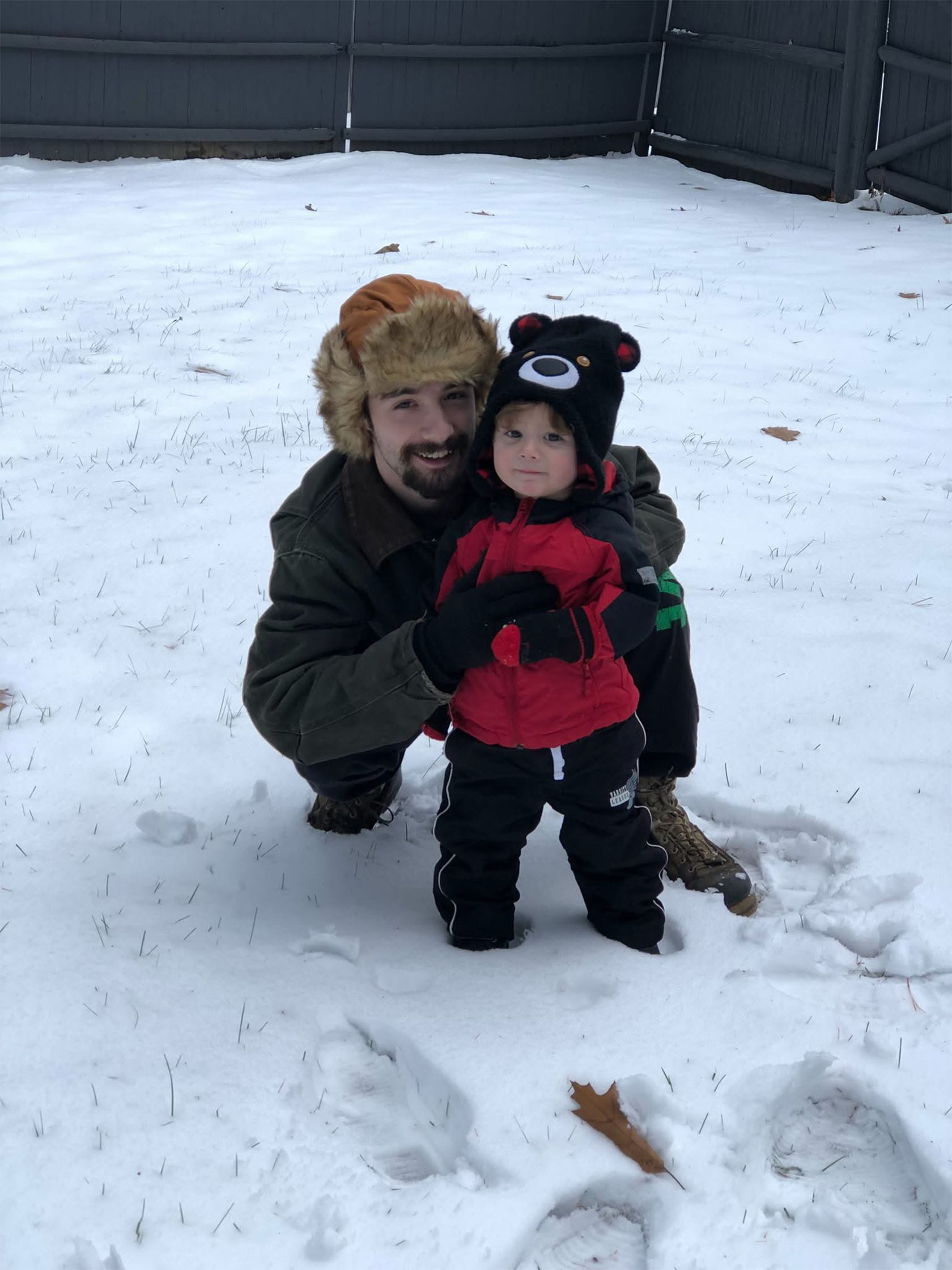 A man wearing a winter hat crouches in the snow beside a young child in a red jacket and bear-themed hat, both bundled up for cold weather in a snowy backyard.