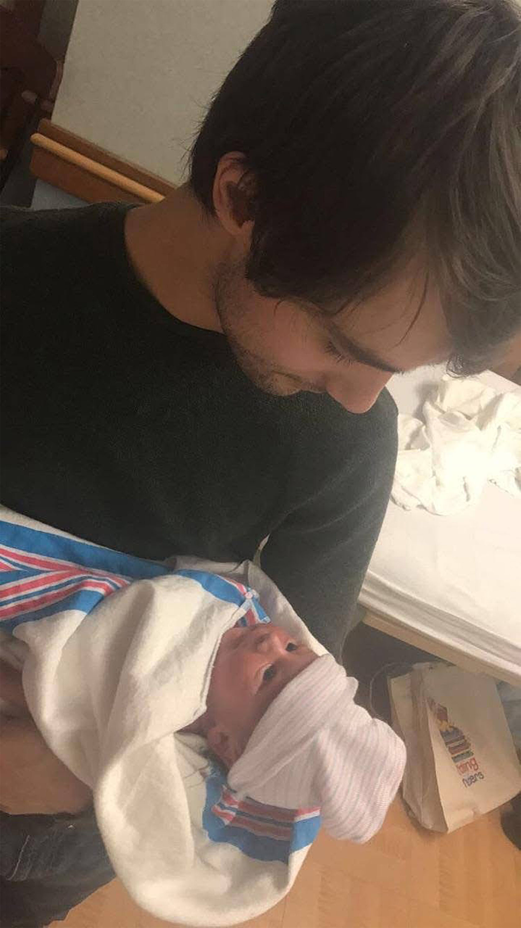 A man wearing a dark sweater holds a newborn baby wrapped in a hospital blanket, looking down at the infant in a softly lit hospital room.