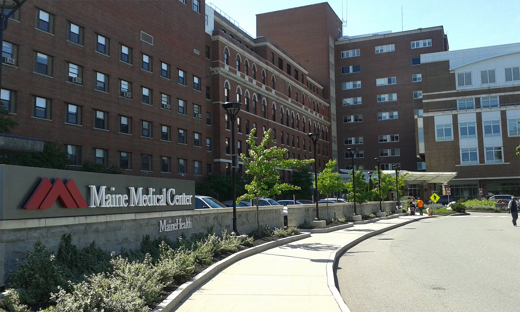 The exterior of Maine Medical Center, a large brick hospital complex with multiple connected buildings, a sign reading “Maine Medical Center MaineHealth,” and a curved sidewalk lined with small trees on a sunny day.