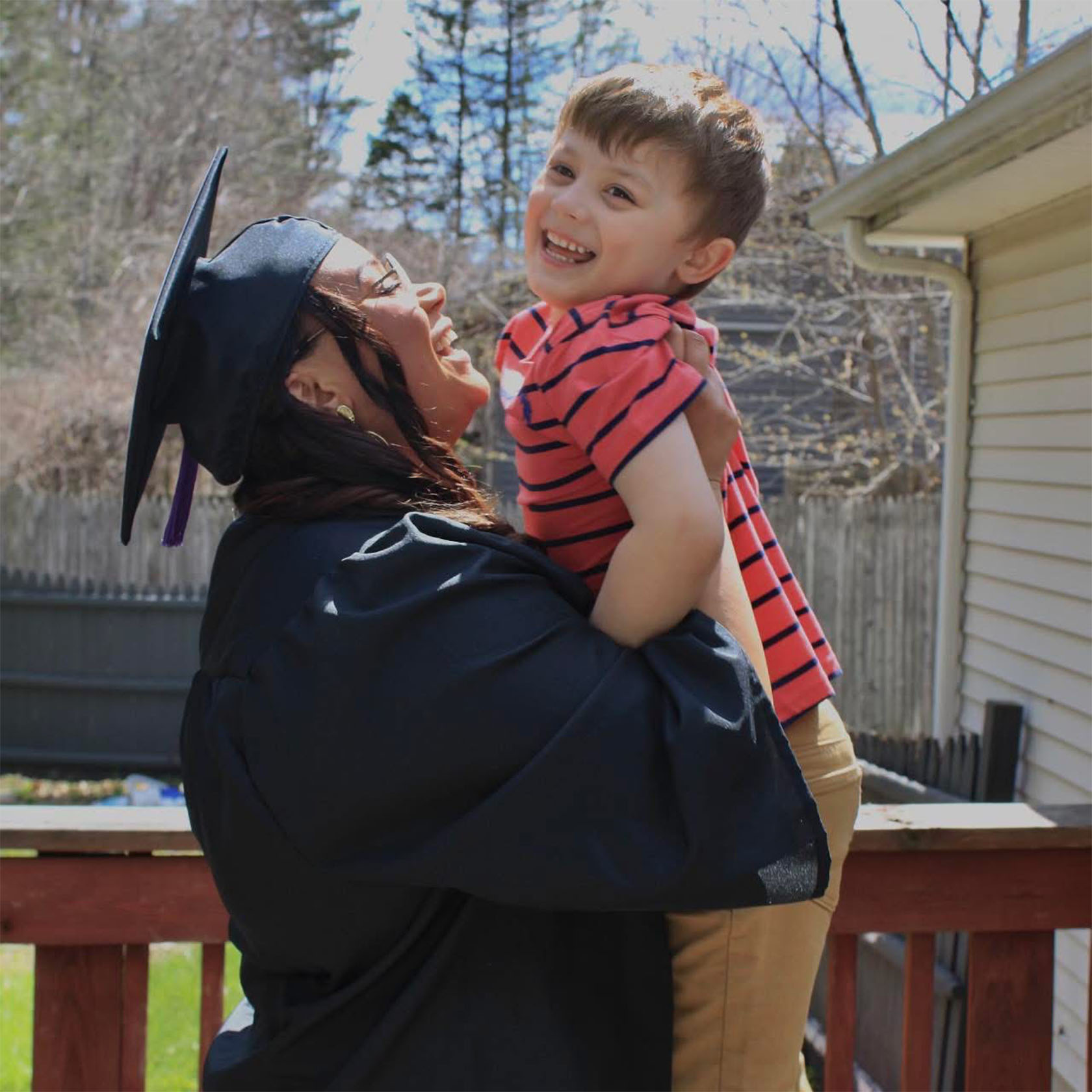 A woman wearing a graduation cap and gown smiles while holding a young child in a red striped shirt on a sunny day outdoors.
