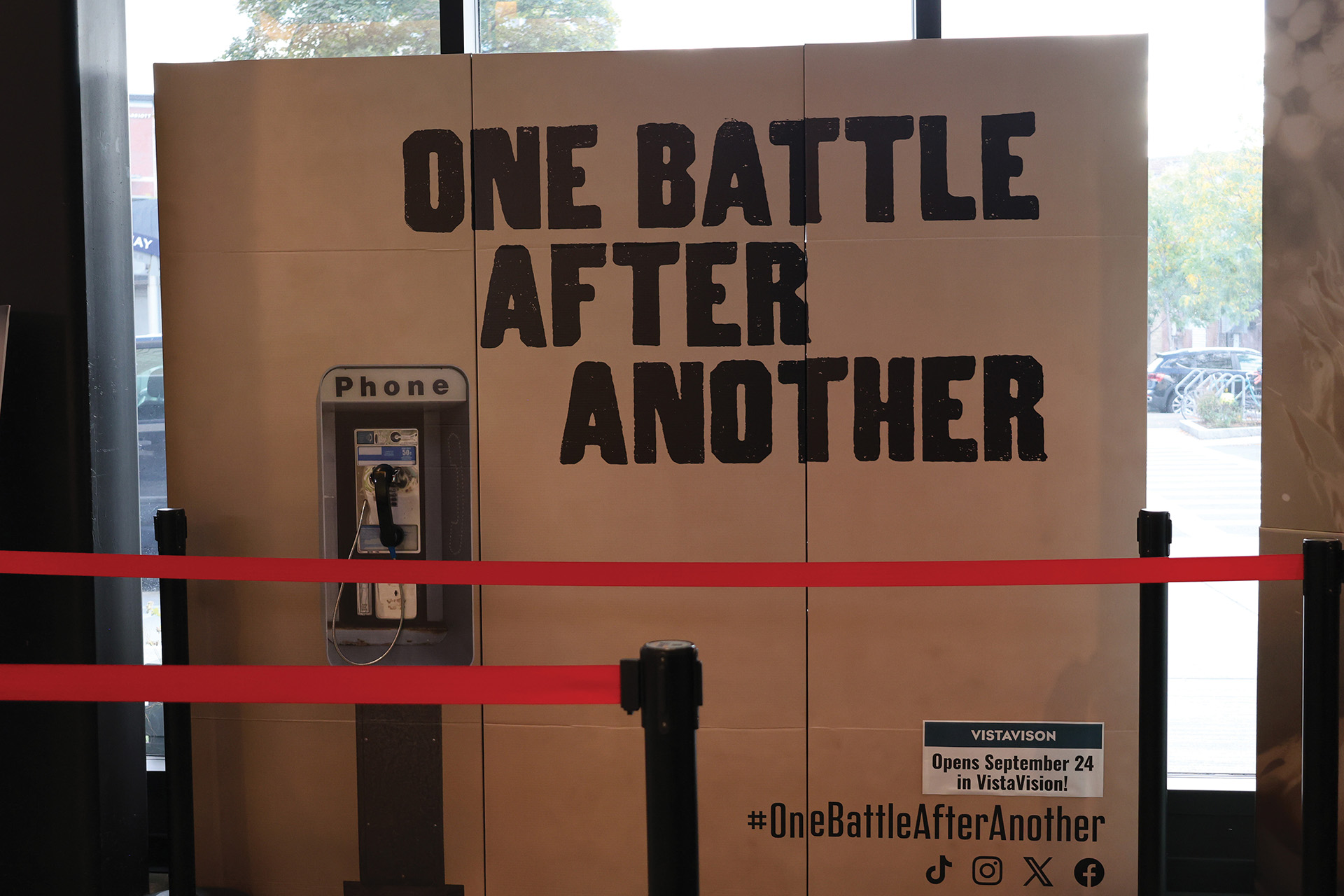 Photo of a large cardboard sign that says One Battle After Another with an attached phone where guests can take pictures to celebreate the films release