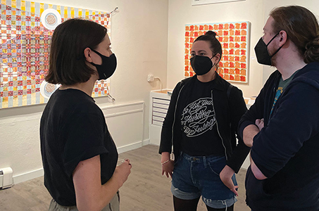 Three people wearing face masks talking in a gallery space with a painting behind them