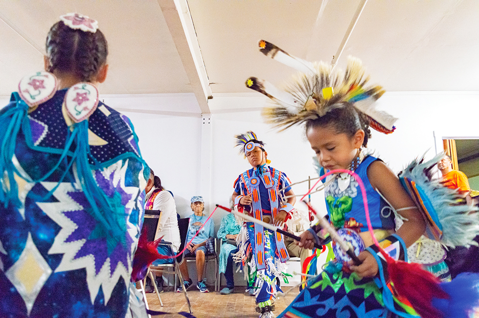 Children in traditional powwow regalia dance around a drum circle.