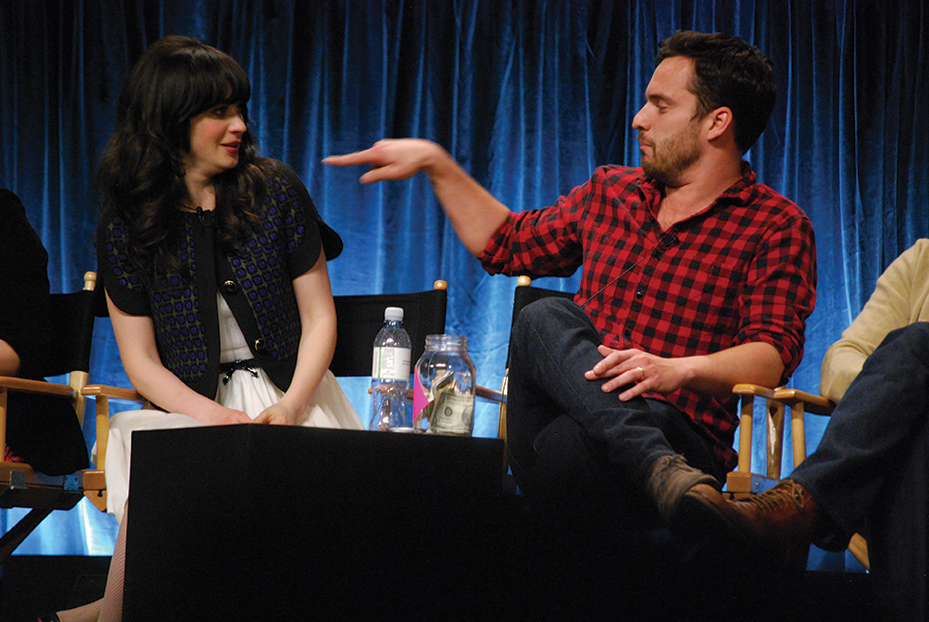 Actor Jake Johnson, wearing a red flannel shirt, points at actress Zooey Deschanel, wearing a white dress and a blue jacket, while sitting on chairs at a panel discussion.