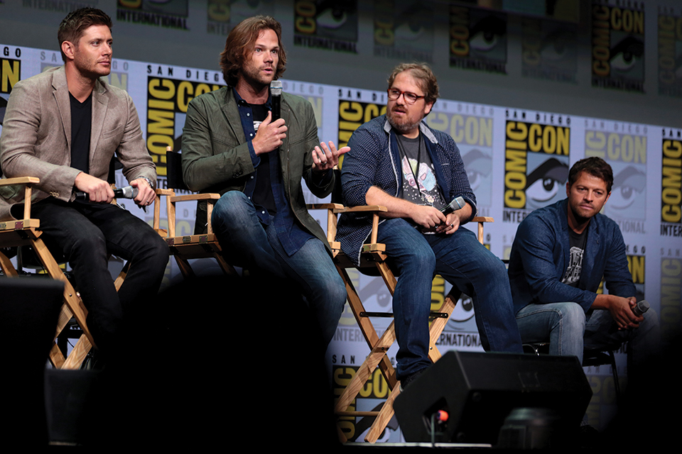 Actors Jensen Ackles, Jared Padalecki and Misha Collins and writer Andrew Dabb sit in chairs on a stage during a panel discussion.