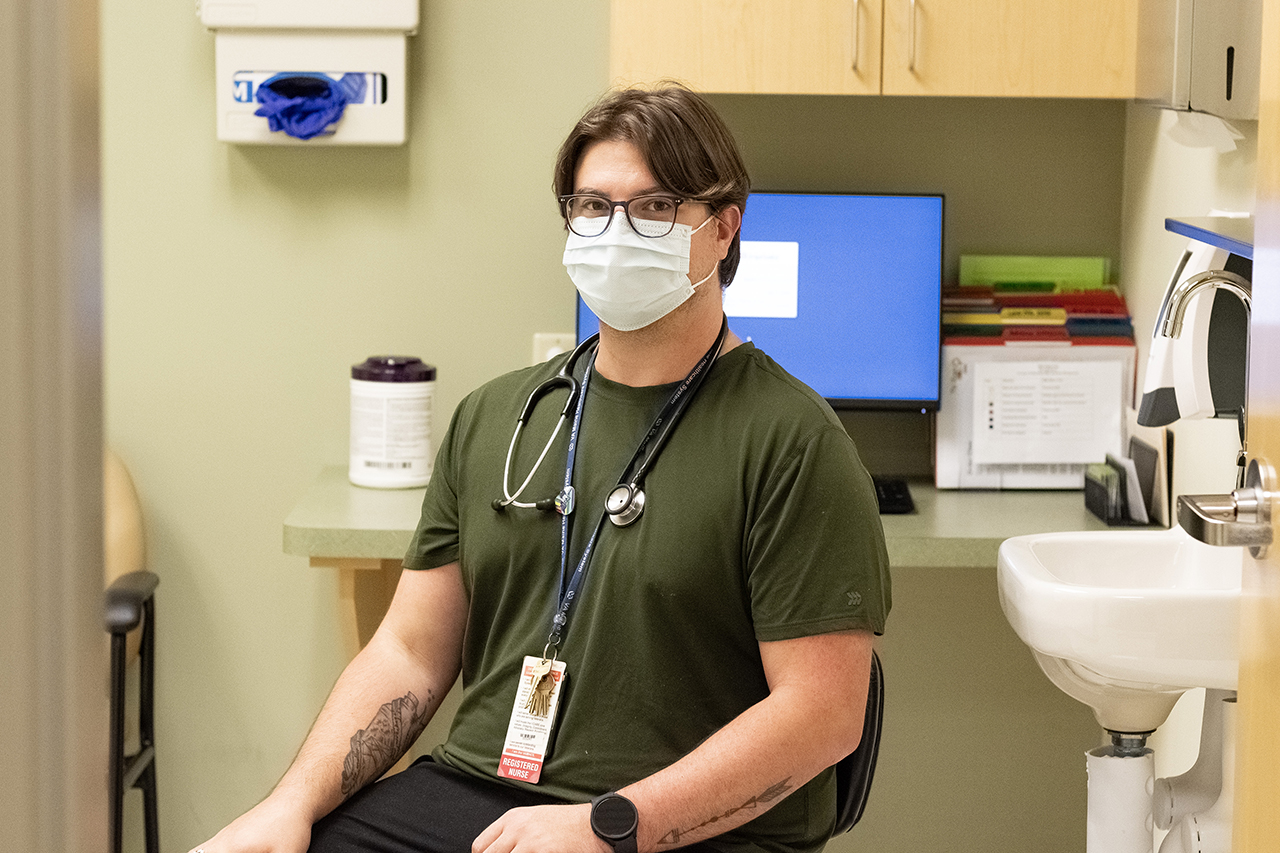 pictured: Nick Durand, sitting down looking at the camera with a mask on. He has tattoos and glasses with longer hair. He is sitting in a patient room surrounded by medical equipment.