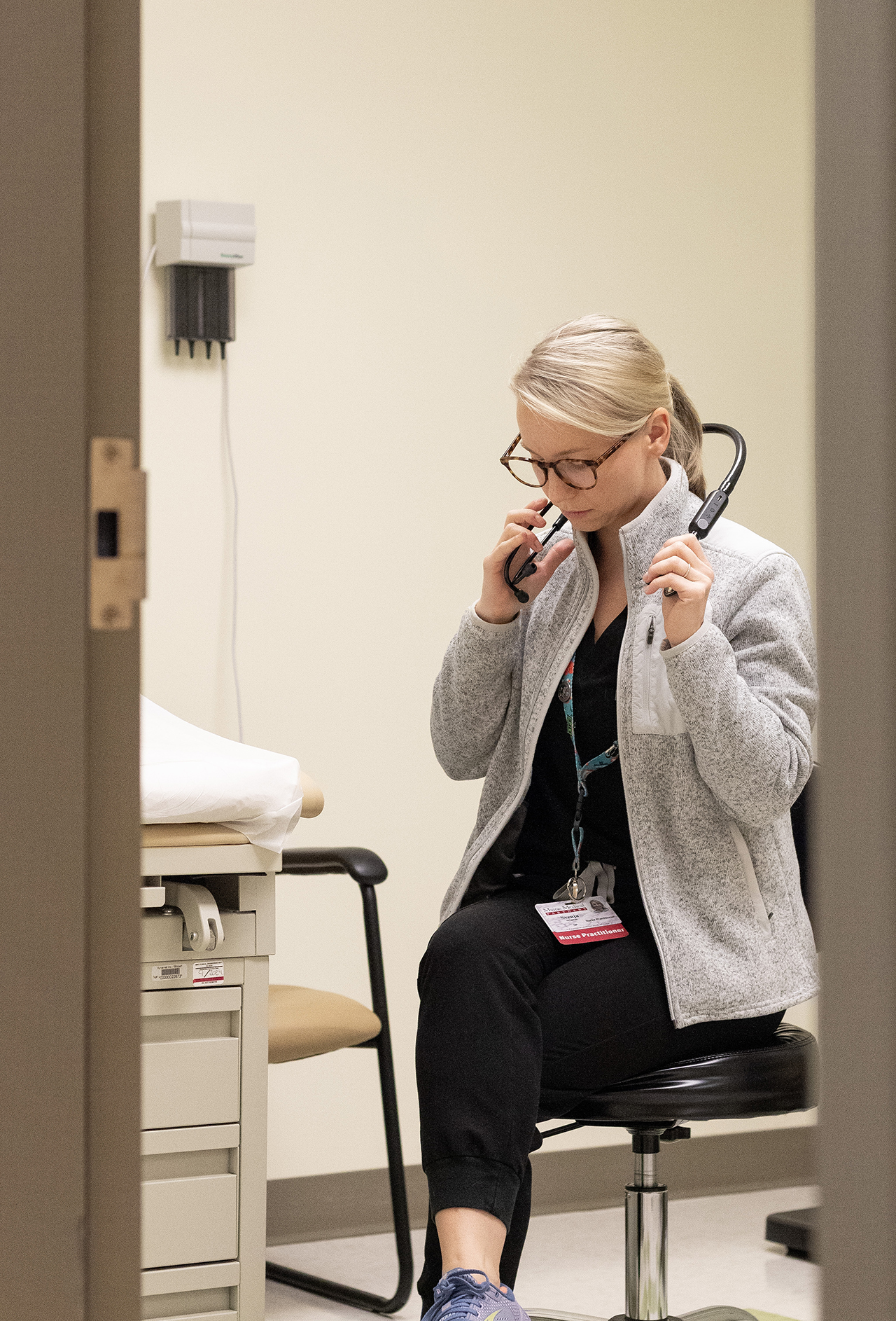 pictured: Svenja Sjoquist, sitting on a stool surrounded by hospital equipment. She has blonde hair in a pony tail and large glasses. She is speaking to a patient that cannot be seen.